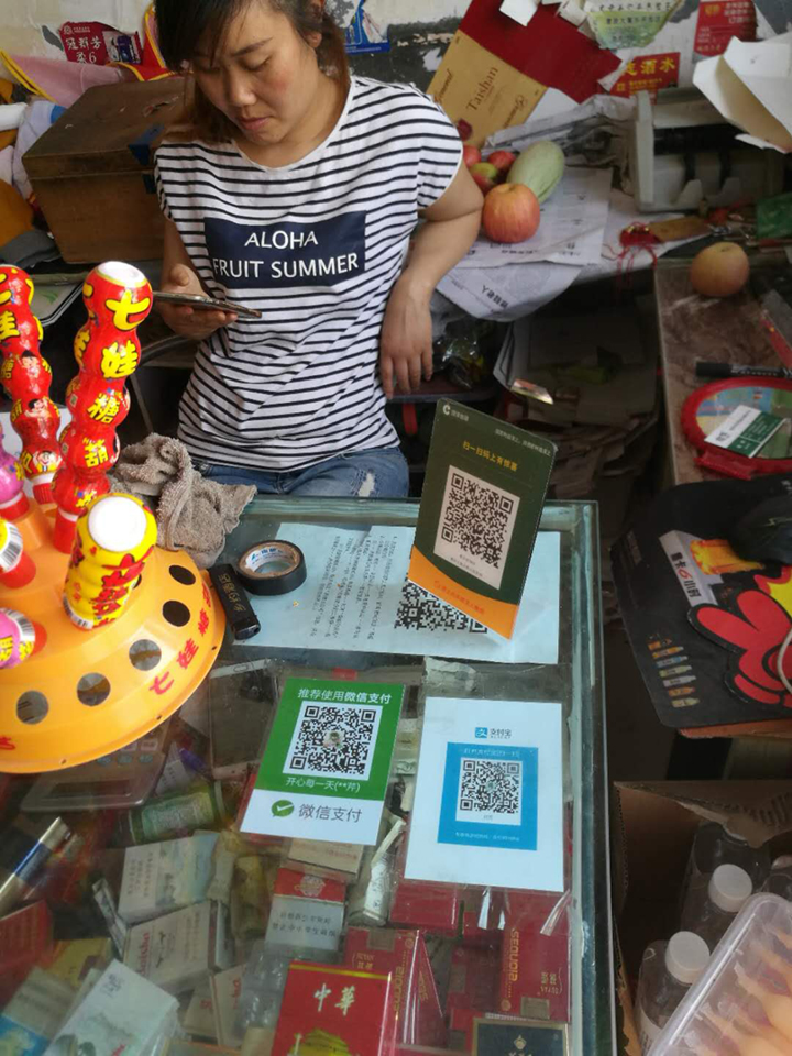 A shopkeeper stands behind a glass counter displaying QR-code payment cards: a green WeChat Pay card on the left and a blue Alipay card on the right.
