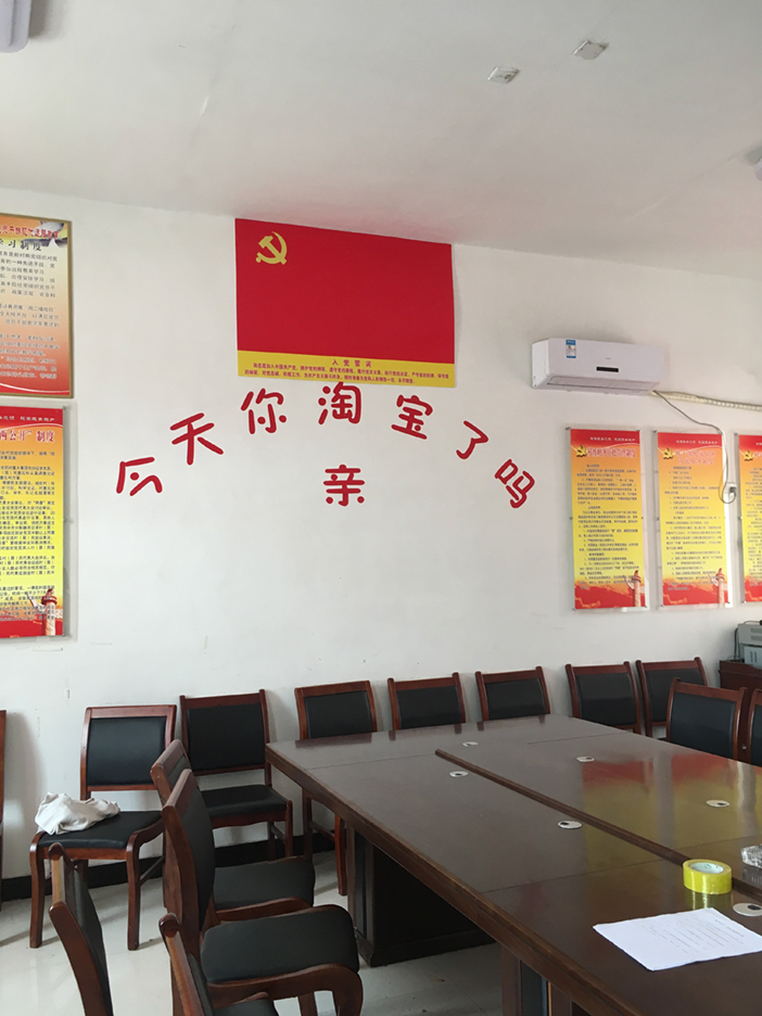 A village government meeting room has a long table and rows of chairs. Beneath a red Communist Party flag, large red Chinese characters on the wall ask, ‘Have you Taobao-ed today?’