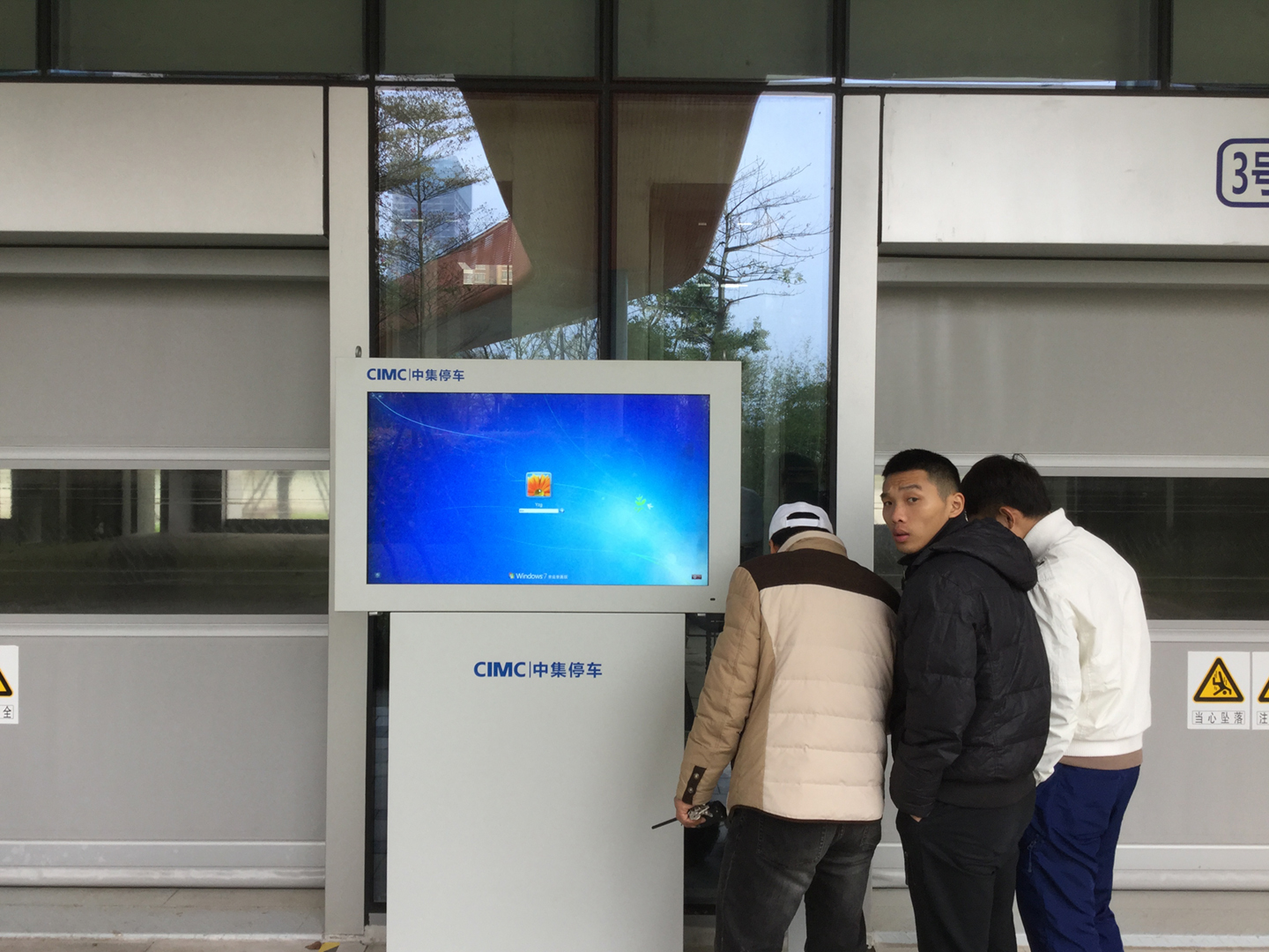 Three workers gather around an automated parking-garage terminal displaying a Windows login screen, beside large metal garage doors.