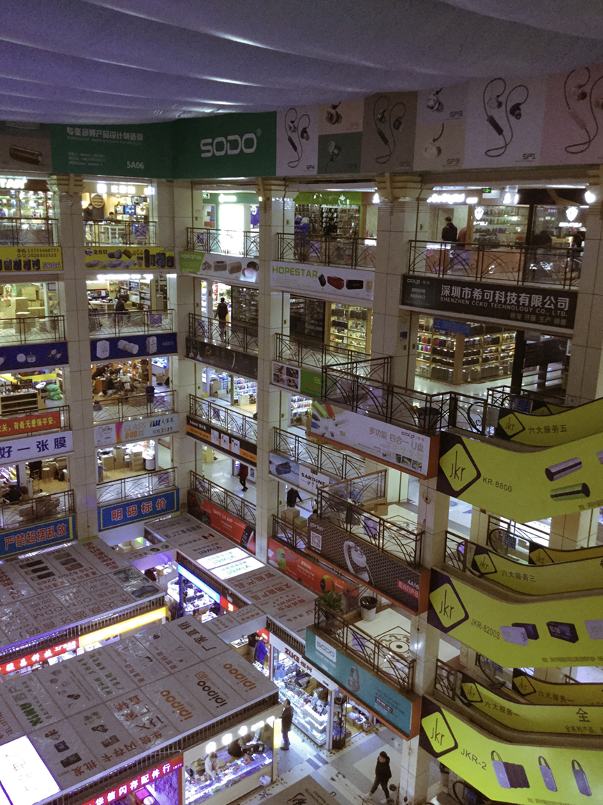 Several storeys of brightly lit electronics stalls surround an open atrium in Huaqiangbei’s market. Shop signs and packaged goods crowd the railings and storefronts.