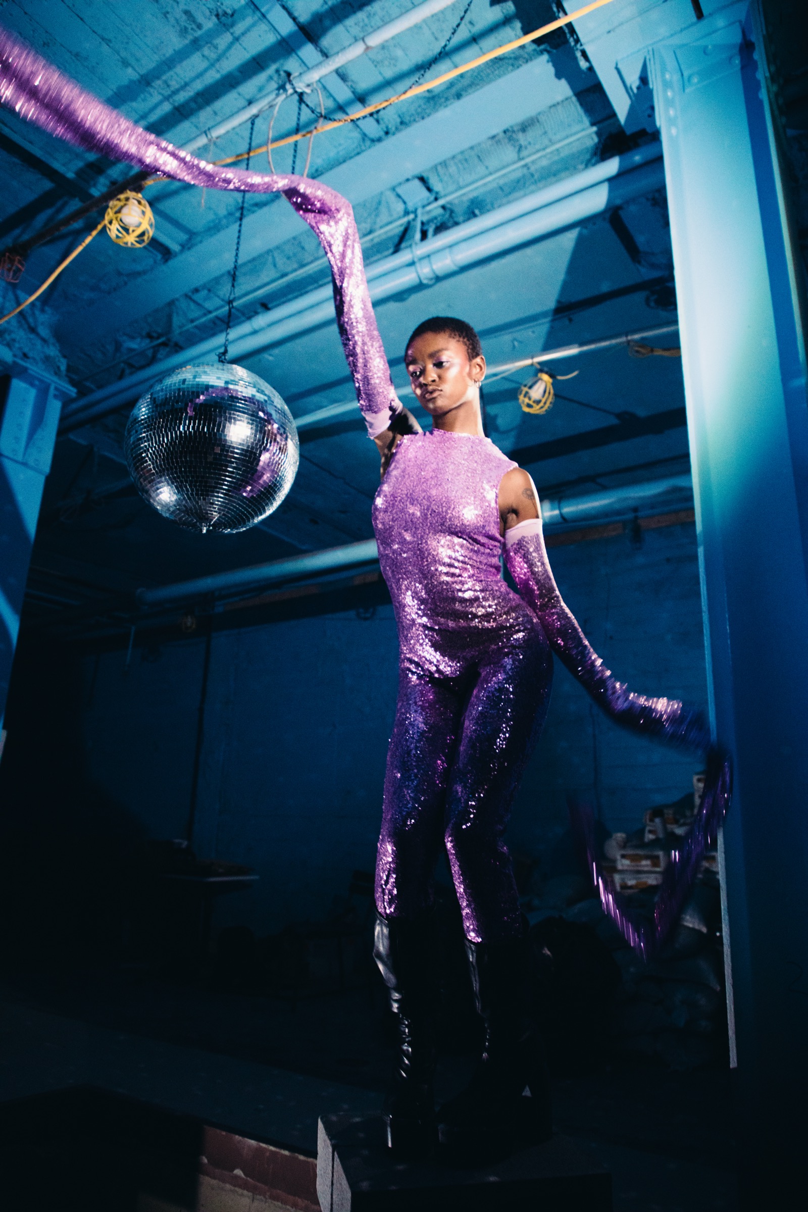 The same model and outfit from the previous photo is displayed in a different
     pose, showing another angle to the garment's draping; the model holds the chest-level
     purple pleated fabric like a fan. The background is dark grey.