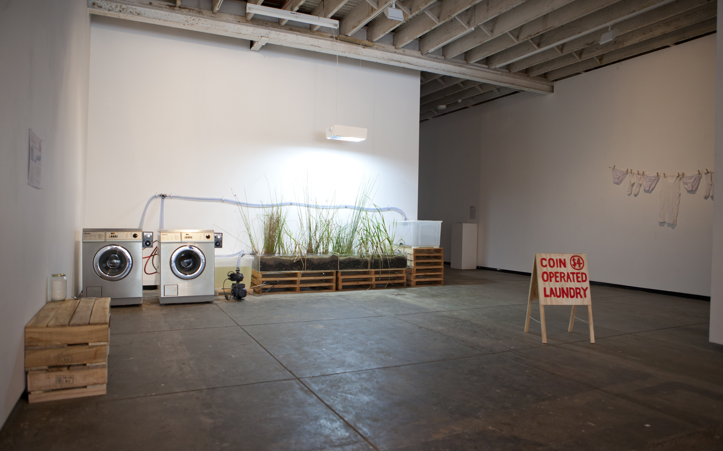 Photo of an installation with two washers wired to a planter, with a sandwich board reading "Coin operated laundry" in the foreground.