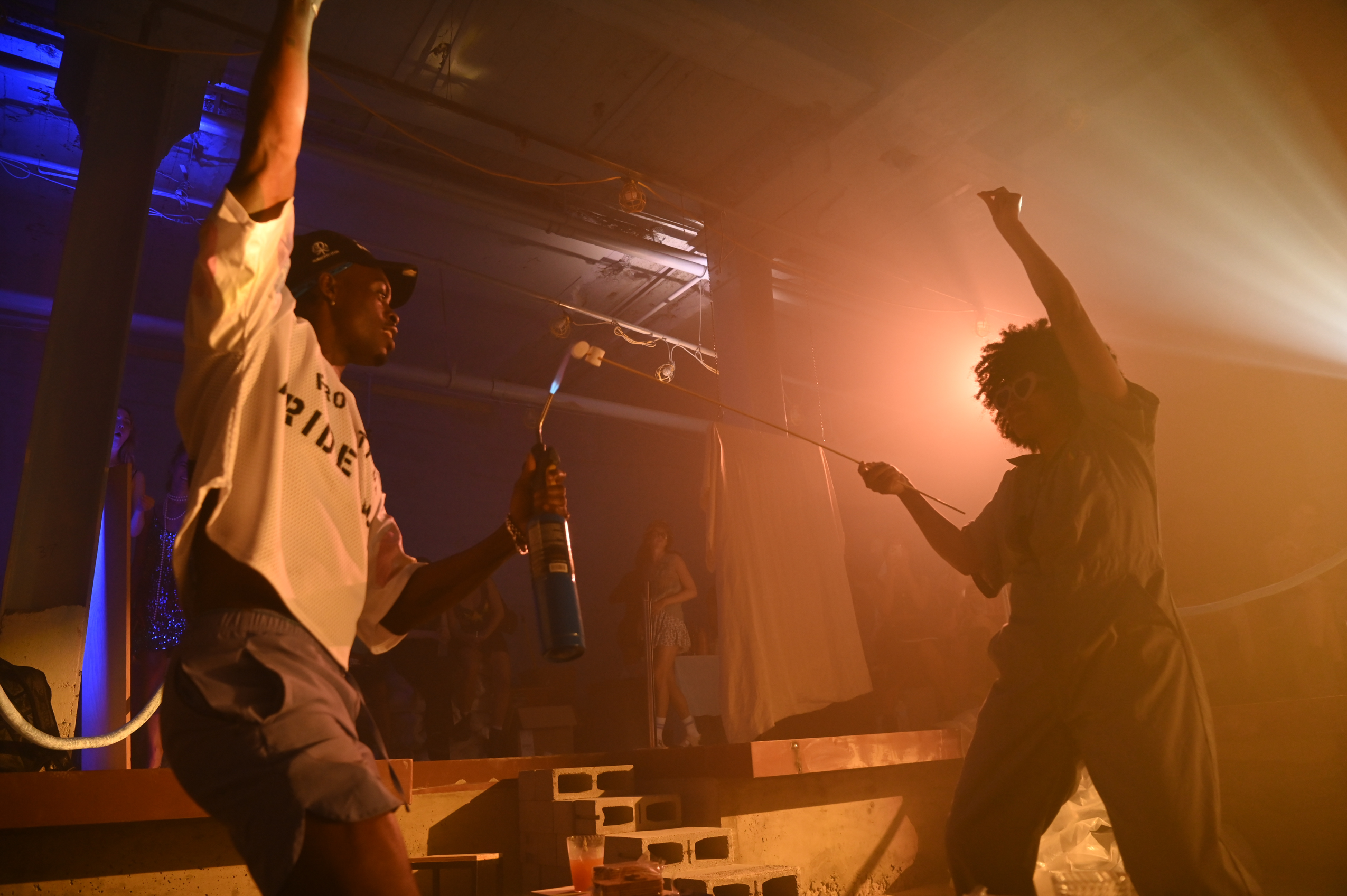 Two people dance with raised arms in orange haze beside a low stage. The person on the left holds a microphone; blue light illuminates the ceiling behind them.