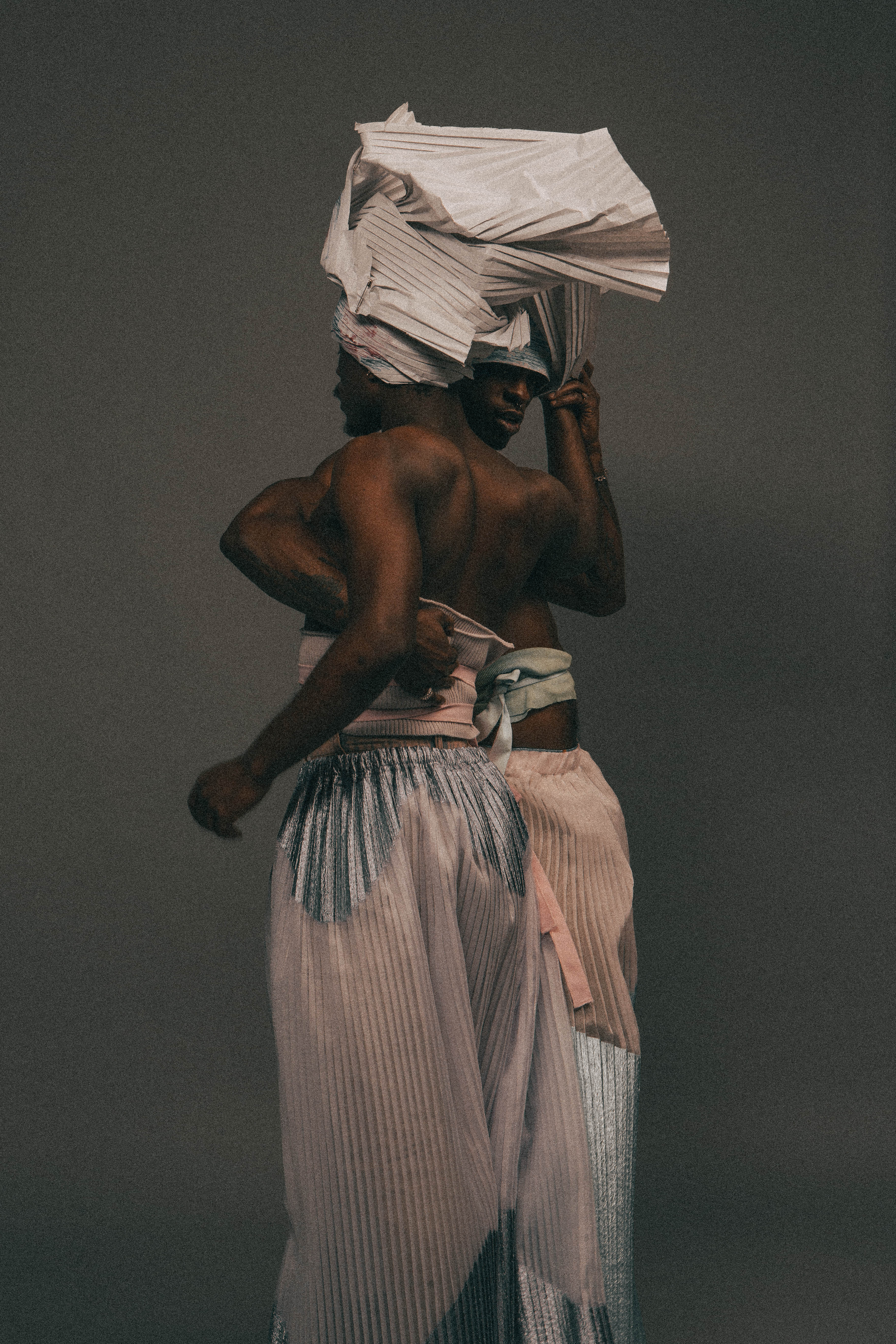 Two models half-embrace each other, with a pleated paper head wrap that encompasses the space above both of their heads. The model on the left wears a ribbed lavender and pink tube top with wide pleated pants, with shimmery blue fabric at the top and white pleated fabric for the rest. The other model wears a blue garment tied at the waist, and pleated wide pants with the same two fabrics, but with the shimmery blue fabric at the bottom half of the pants.