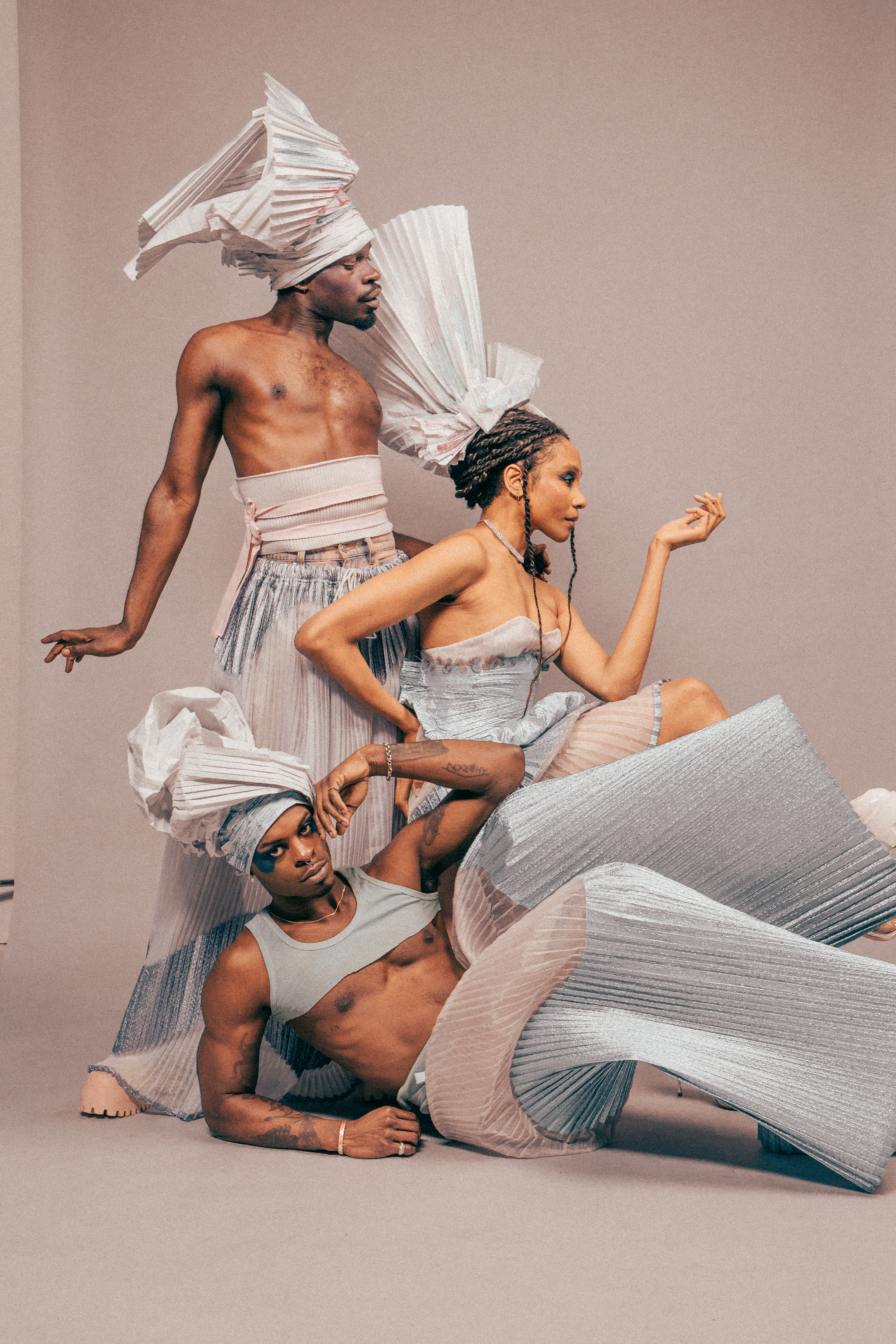  Three models pose together: one on the left of the photo is standing up, with their wrist flared out to one side; a woman to their right sits down facing right, with one hand on her hip and one resting on her thigh; the last model poses in the bottom half of the photo, lying on their side balanced on their arm. Each of them are wearing different pleated paper headpieces with complex shapes, as well as some jewelry, and pale pink and blue garments made of alternating fabrics, mostly pleated or ribbed. The model in repose is looking pointedly and directly at the camera, while the others gaze to the side.