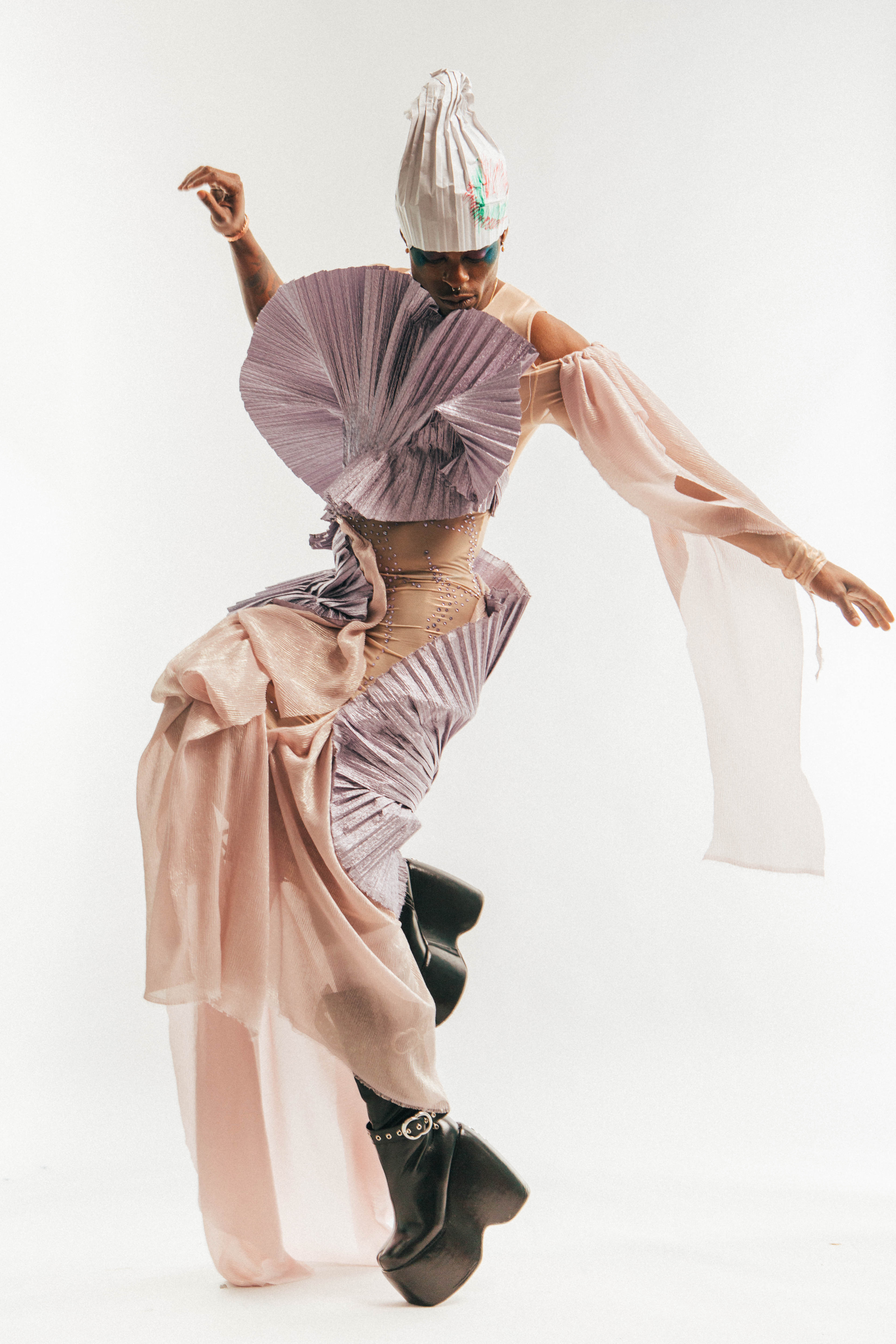  A Black model with their arms splayed out, one leg up, in a dancing posture. They are wearing a white draped headpiece with red and green drawings on the side, a shimmery dress with lavender pleating on the chest, diaphanous, flowy sheer pink fabric on the arms; these two fabrics flow together through the bottom half of the dress. The bodice is a beige color beaded with rhinestones, which shows through in the stomach area. The model is wearing earrings and has a septum piercing, and they wear black platform boots, against a white backdrop.