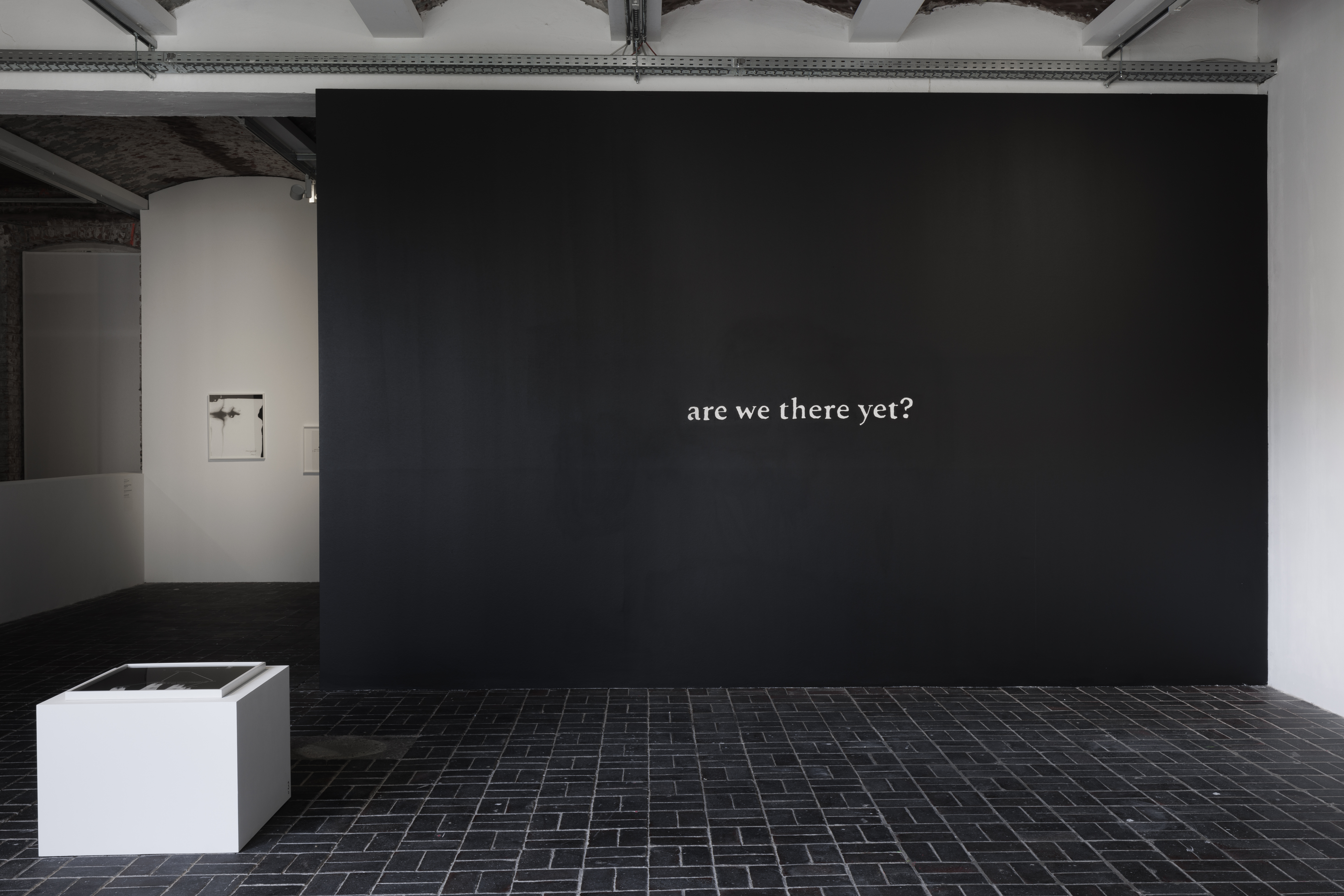 The words ‘are we there yet?’ appear in small white letters at the centre of a large black gallery wall. A low white display case stands in front of a dark tiled floor.