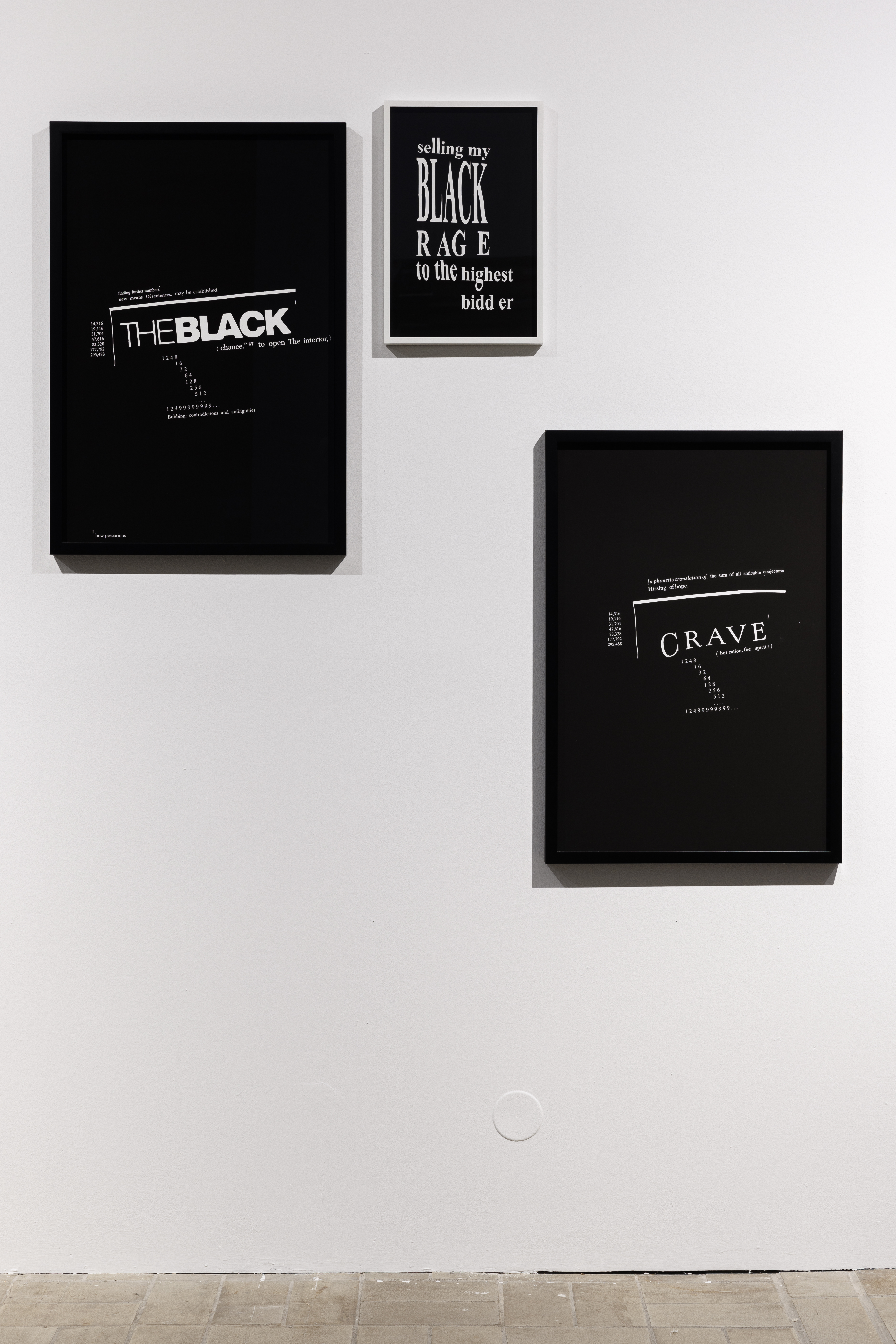 Three differently sized black prints hang on a white wall. Their scattered white typography includes ‘THE BLACK,’ ‘BLACK RAGE,’ and ‘CRAVE.’