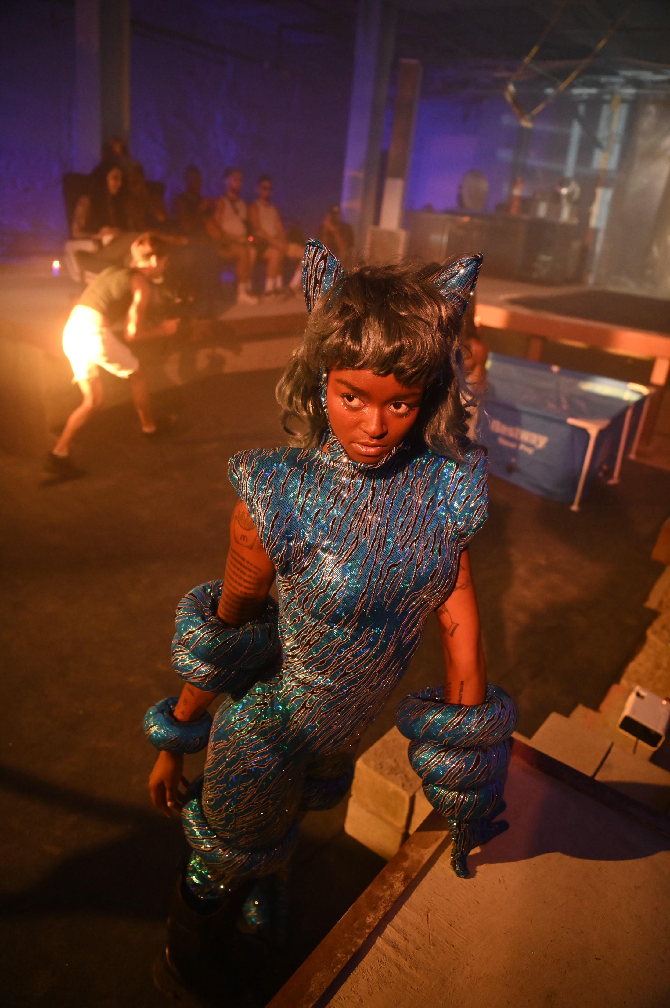 A performer in a textured blue costume, padded gloves, and pointed ear-like accessories looks toward the camera. A fire performer and a portable pool are visible behind them in the dim venue.