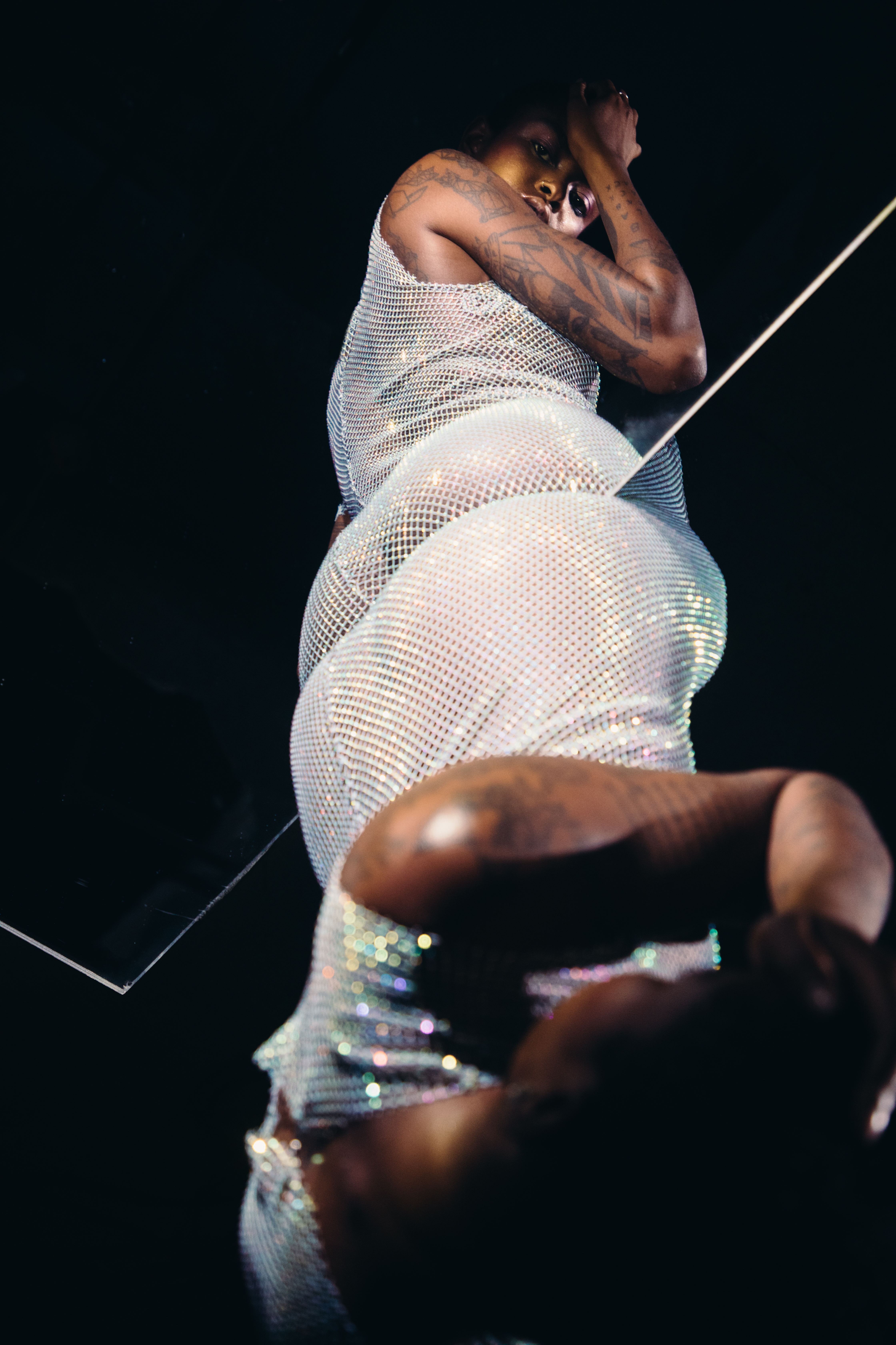 A Black model spins in a black and white pleated head wrap, with a blue, white,
     and silver-grey pleated strapless shirt tied with some ribbons. Translucent light
     green-blue fabric fans out to the model's right. The skirt of their outfit is made
     of a shimmery light green fabric with smaller pleats, and it drapes across their
     legs, against a white backdrop.